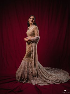 A bride in a shimmering champagne mermaid gown with feathered sleeves poses against a rich red velvet backdrop.