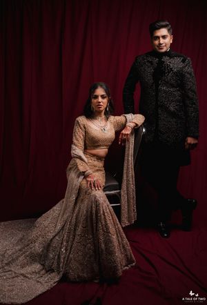 Couple in elegant Indian wedding attire, woman in gold lehenga seated, man in black sherwani standing against red backdrop.