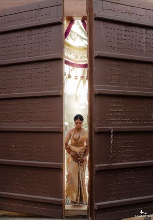 Bride in golden saree standing between large wooden doors at traditional Indian wedding ceremony.