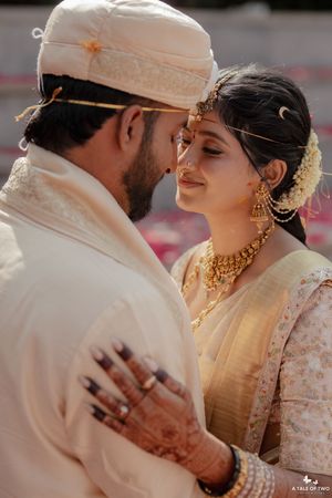 Indian couple in cream wedding attire sharing intimate moment with gold jewelry and mehndi.