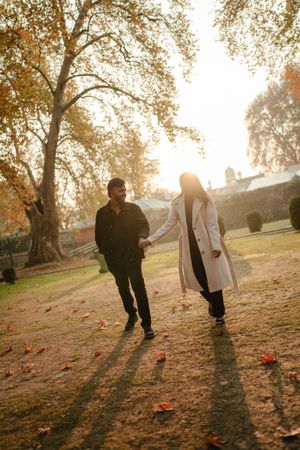 Couple holding hands walking through a sunlit autumn park with golden leaves and trees in warm fall light.
