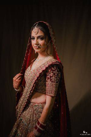 A bride in a red and gold embroidered lehenga with dupatta, adorned with traditional bridal jewelry, poses elegantly.