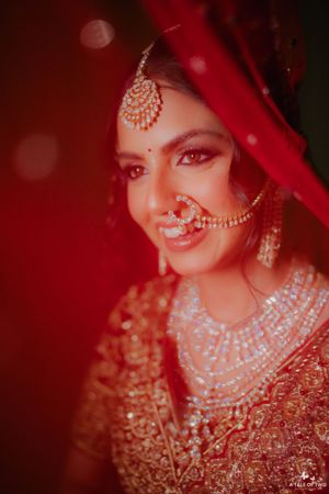 Indian bride in red lehenga with gold maang tikka, nose ring, and layered pearl necklace smiling beautifully.