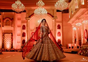 Bride in ornate golden red lehenga twirling at an Indian wedding venue with chandeliers and warm amber lighting.