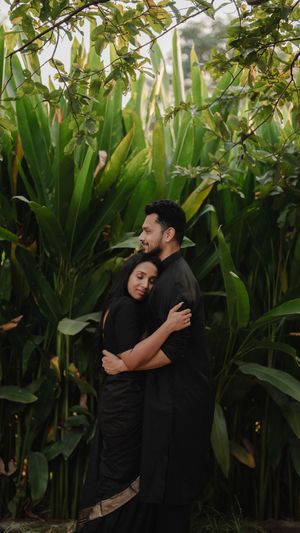 A couple in black clothing embrace tenderly in front of lush tropical foliage and tall green plants.