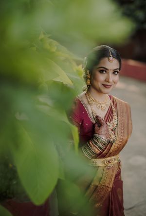 Traditional Indian bridal attire in burgundy saree with gold embroidery and jewelry shown through green foliage.