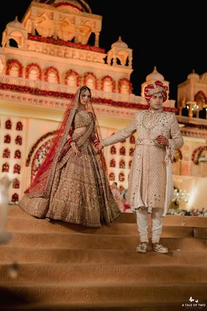 Indian bride in gold lehenga and groom in white sherwani pose on steps before illuminated palace backdrop.