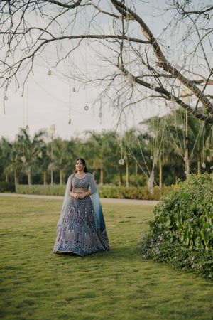Bride in blue lehenga standing on green lawn under bare tree with hanging decorations at outdoor wedding venue.