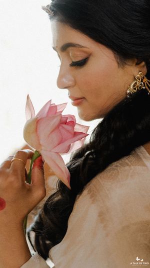 Indian bride in traditional attire smelling a pink lotus flower with eyes closed, wearing gold earrings.