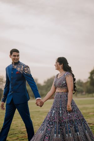 A couple in traditional Indian wedding attire holds hands outdoors, the groom in blue sherwani and bride in embroidered lehenga.