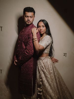 A couple poses in traditional Indian attire, with the woman in a beige lehenga and the man in a burgundy kurta.