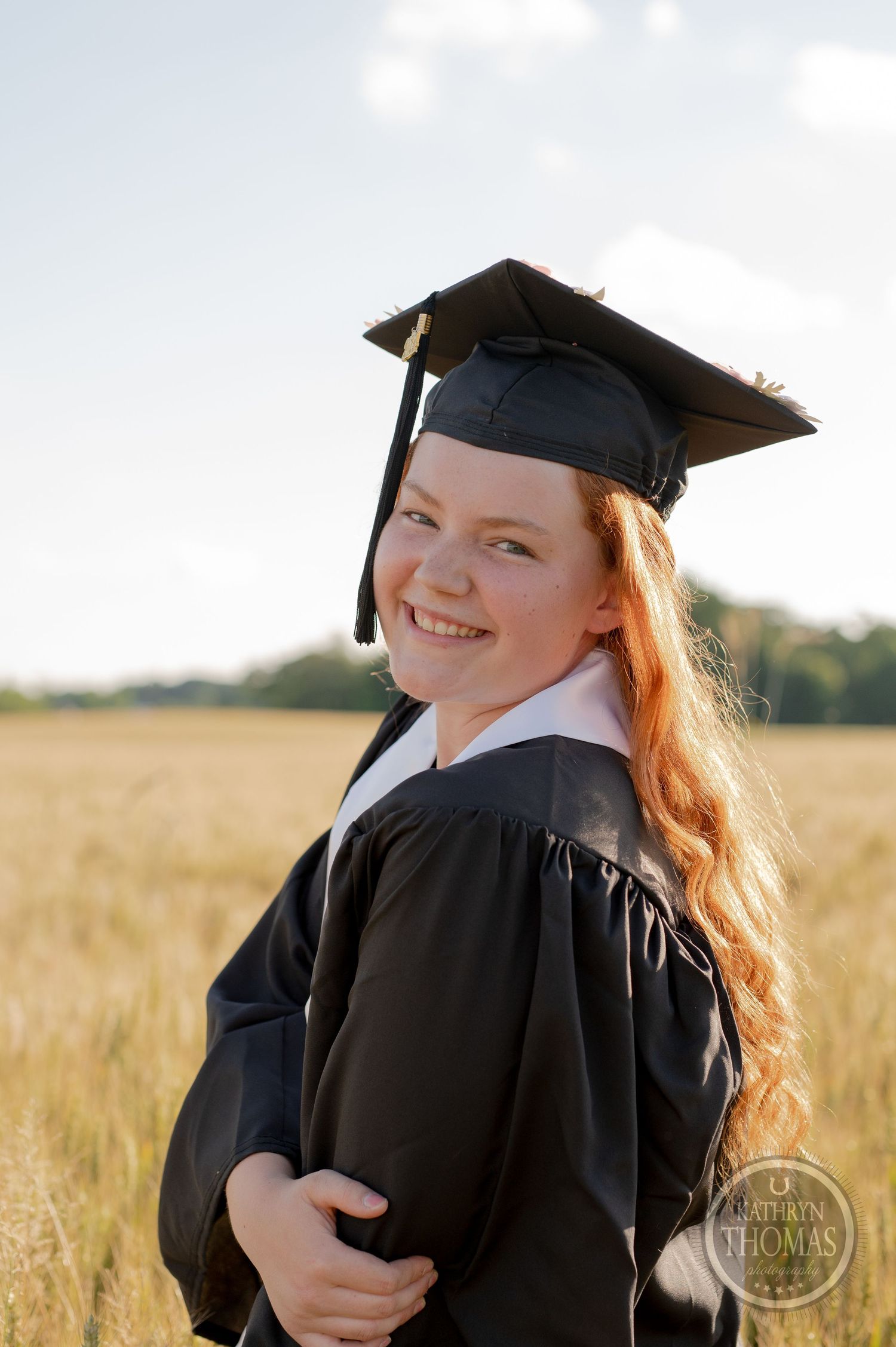 Graduate in full academic regalia poses in wheat field at sunset.