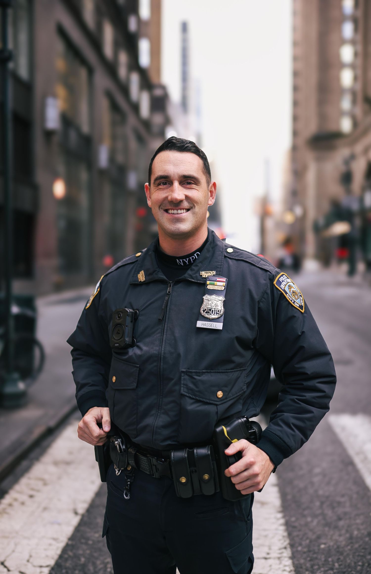 Police officer in dark uniform standing on a city street with buildings visible in the background.