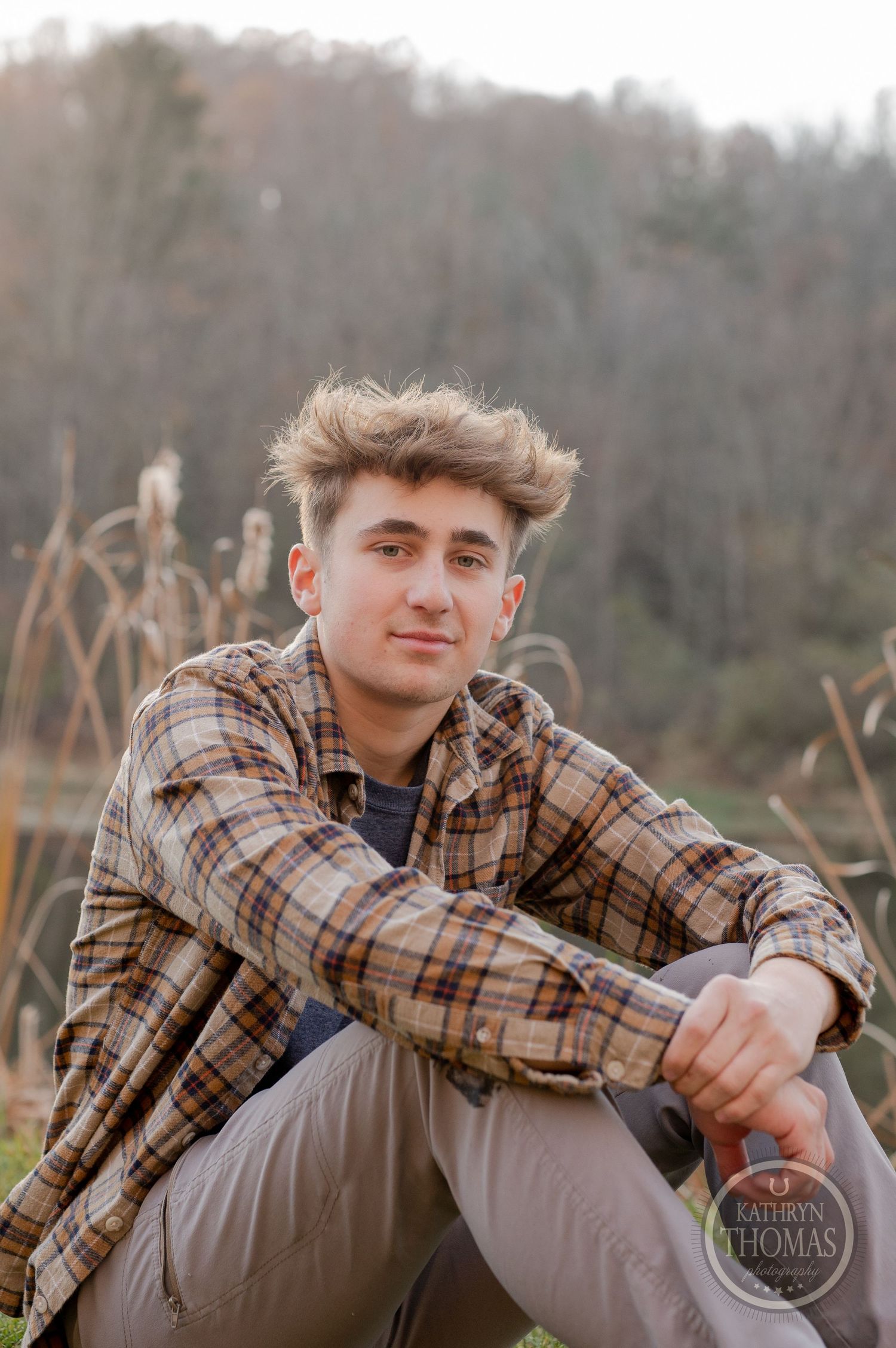 Someone in a brown plaid shirt sits casually on grass near cattails and a lake.