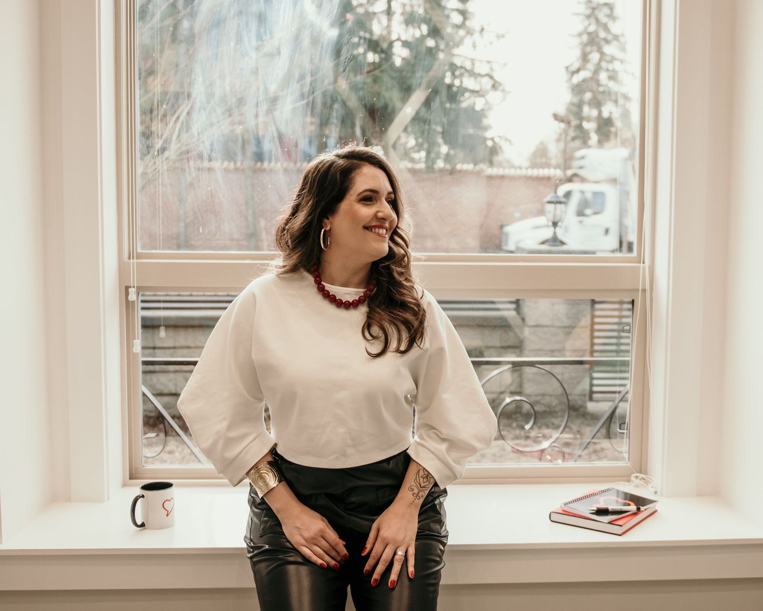 Woman in beige sweater smiling by window in casual lifestyle portrait.