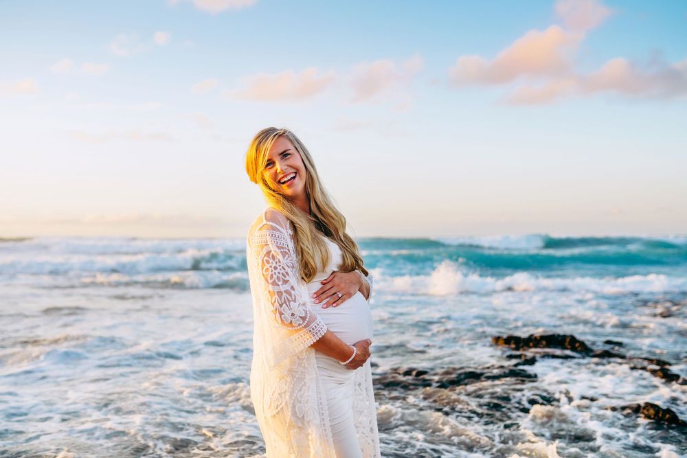 kona maternity photography big island hawaii. Couple in white clothing embracing on rocky coastline with waves splashing at sunset.