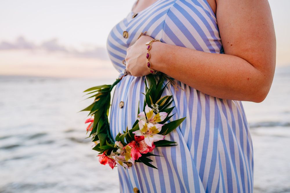 haku lei maternity photos maui hawaii. woman in blue striped dress holding tropical flowers against ocean backdrop at sunset.