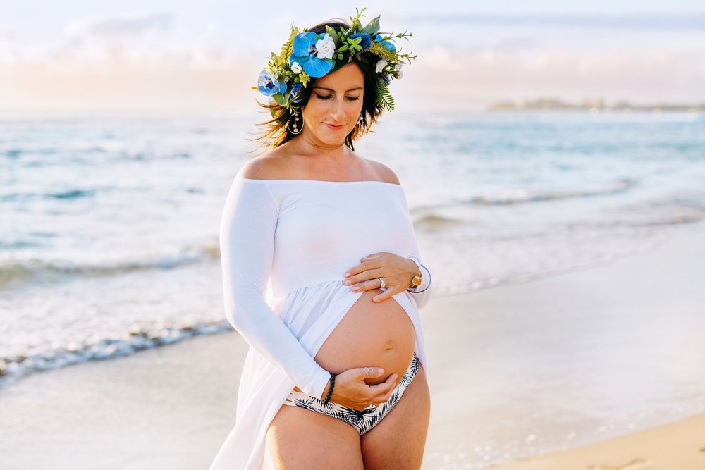 haku lei maternity photos in maui hawaii.Expectant mother in white dress and blue flower crown poses gracefully on tropical beach at sunset.
