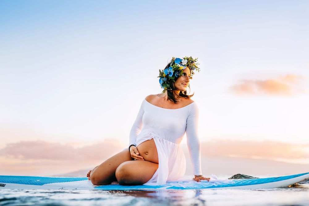 ocean surfboard maternity photos in maui. Maternity beach portraits with flowing white dress against ocean waves and soft sunlight.