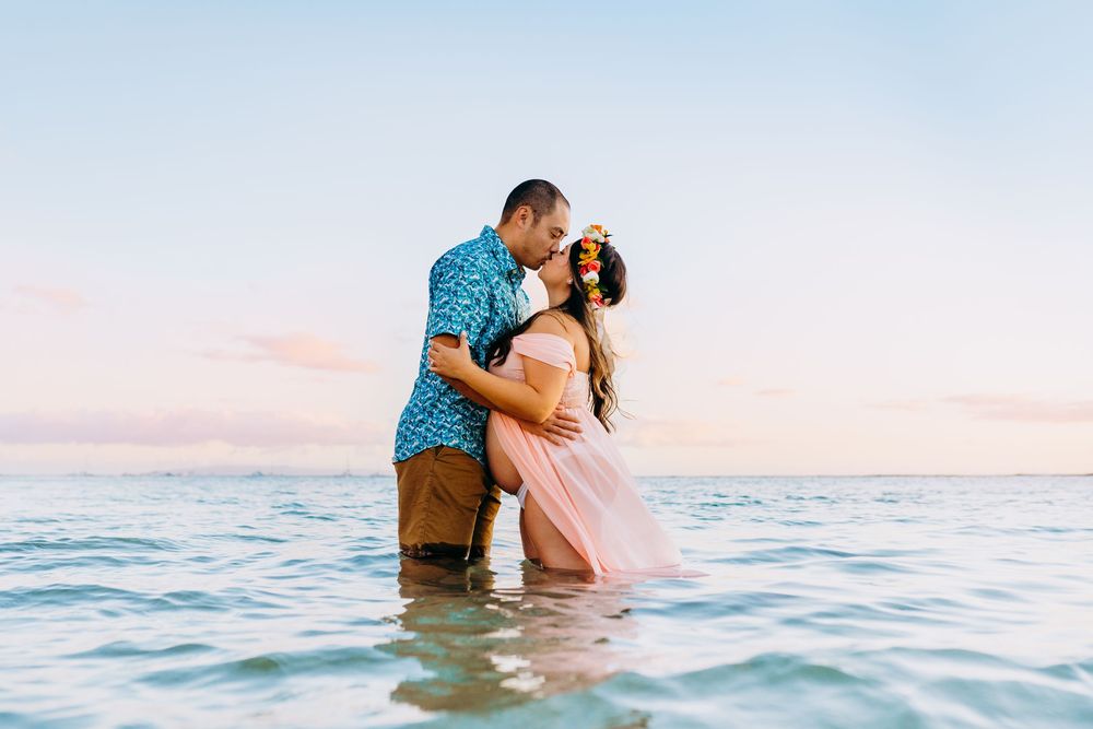 haku lei ocean maternity photos. Couple sharing romantic moment in ocean water during sunset with pink sky.