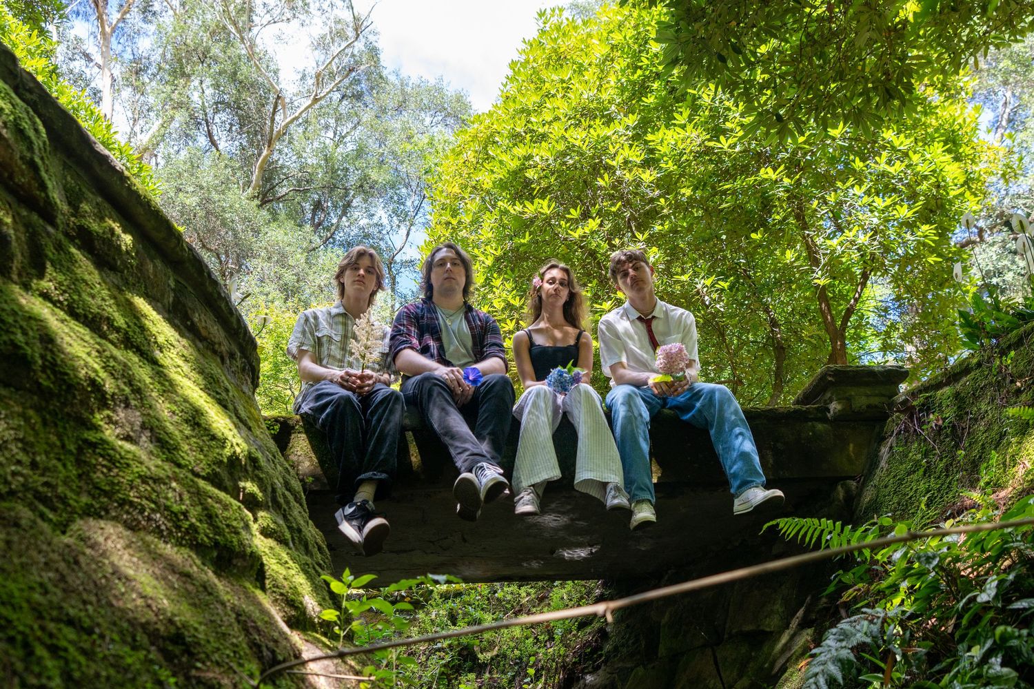 Four band members sit together on mossy rocks surrounded by lush green forest trees.