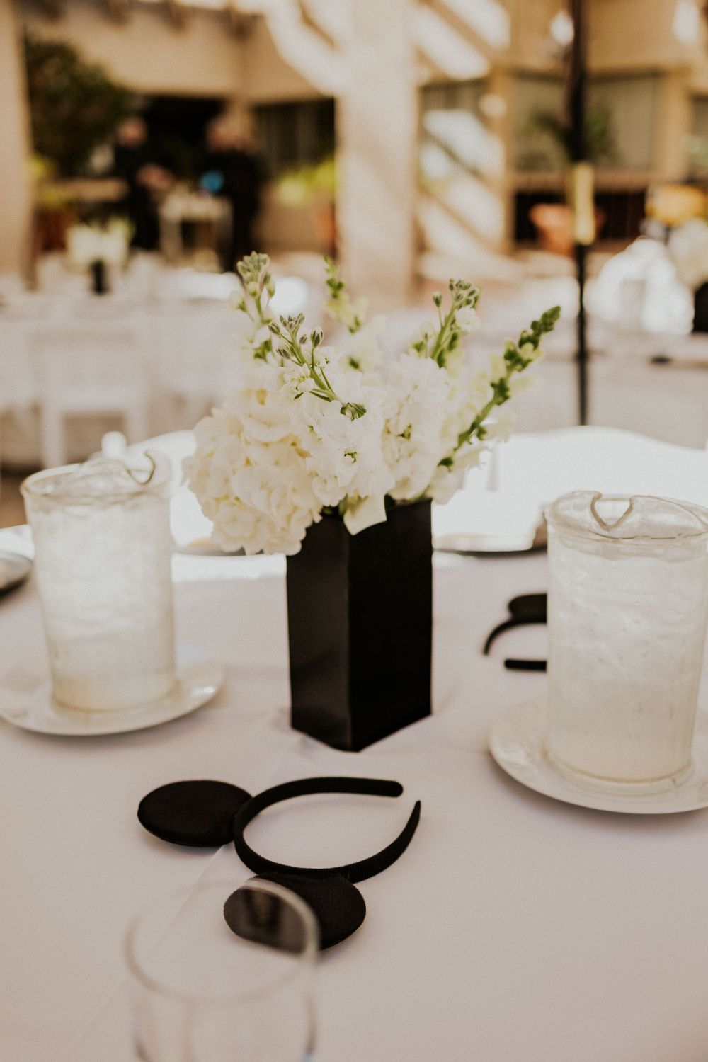 Elegant table setting with white flowers in black vase and Mickey Mouse ears on white tablecloth.