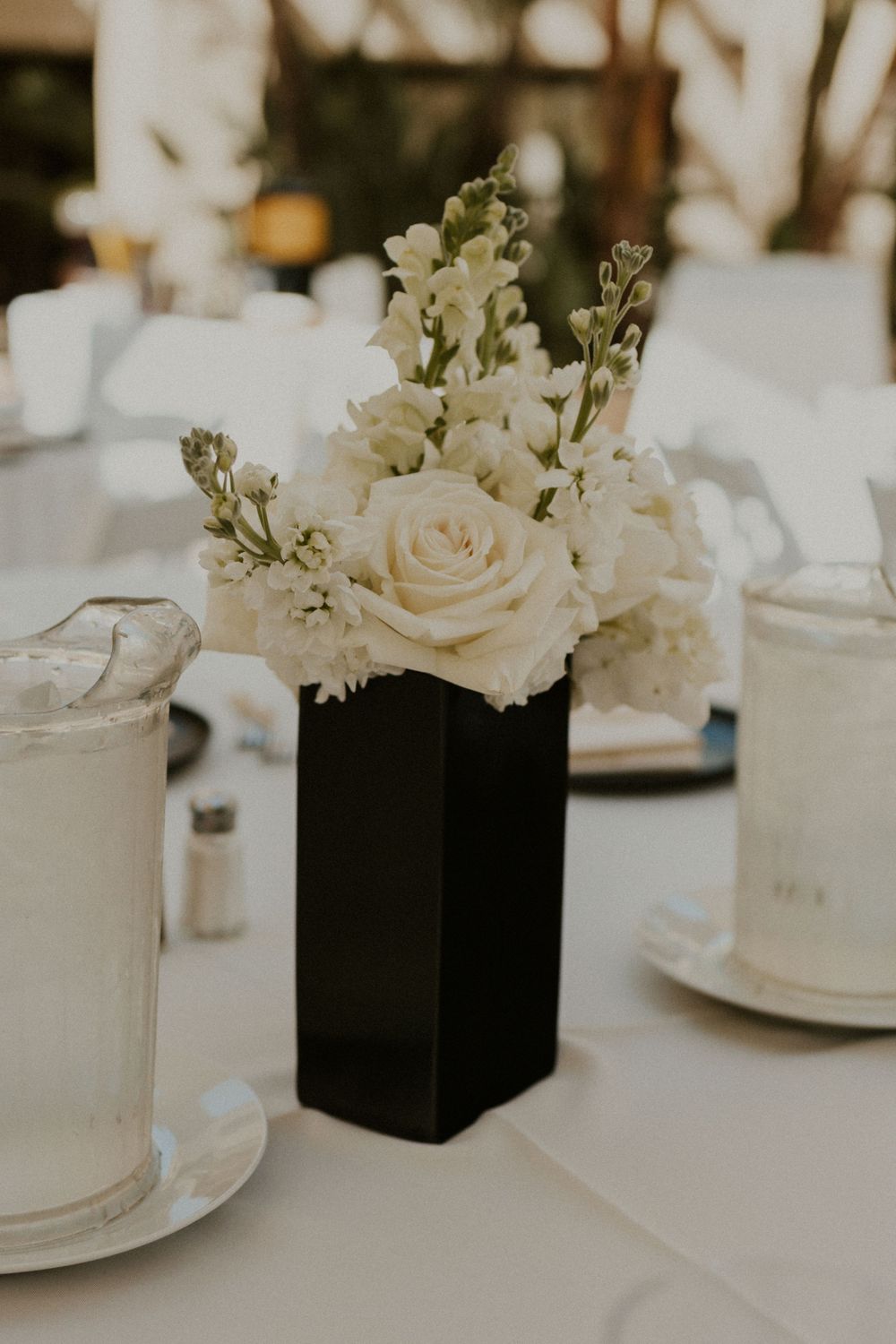 White roses and flowers arranged in black rectangular vase on elegant table setting.
