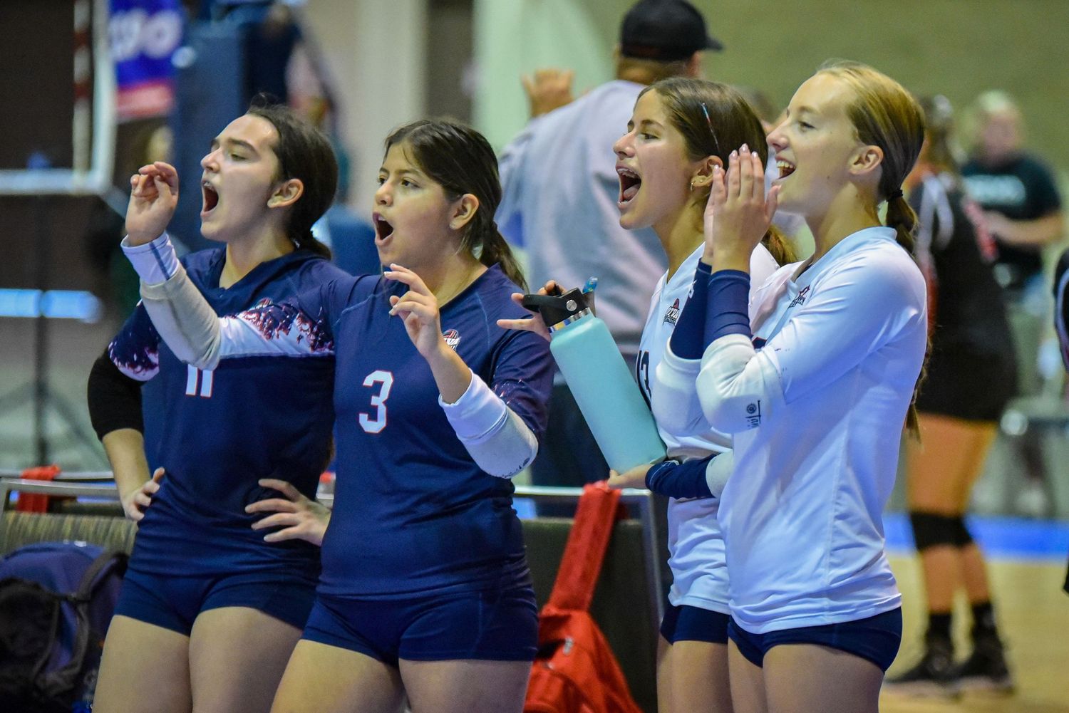 Women's volleyball team celebrates in huddle formation on blue court.