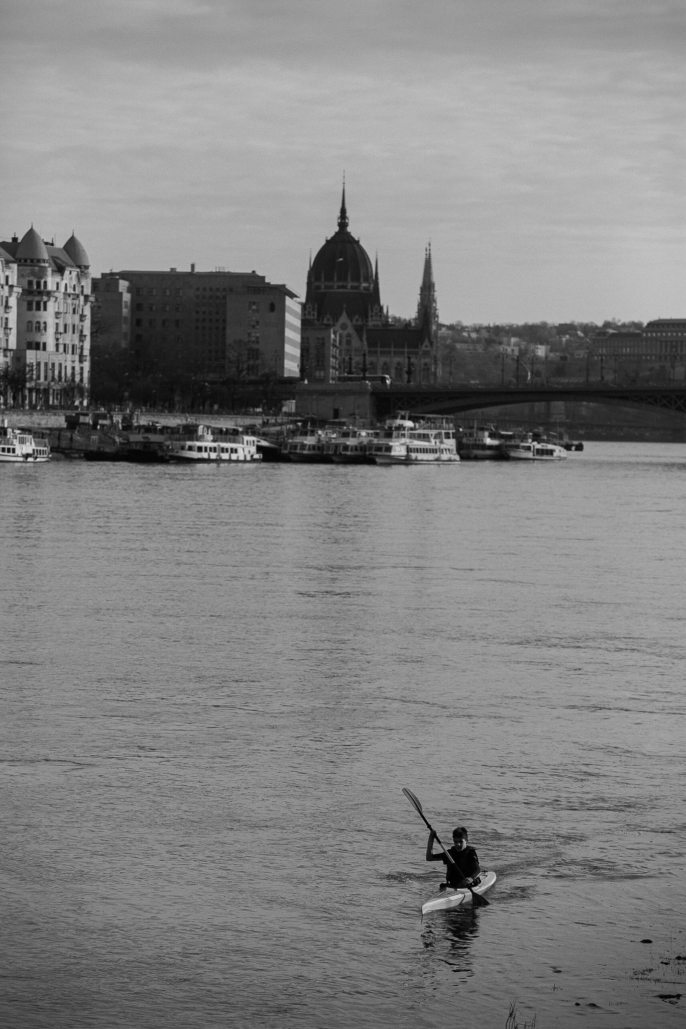A lone kayaker paddles on the Danube River with Budapest's historic Parliament building in the background.