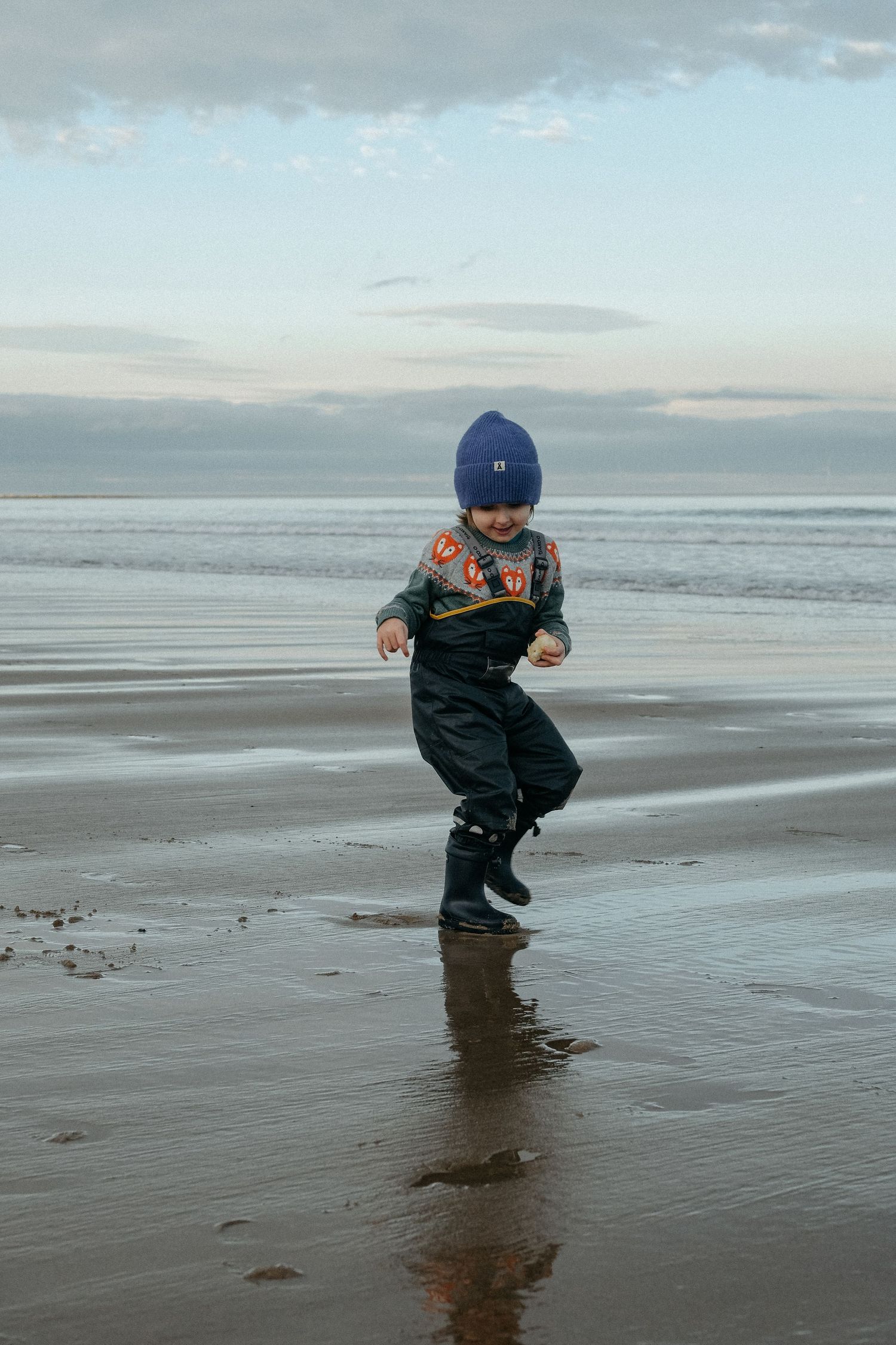 A child in winter clothing playing on a wet sandy beach at dusk.