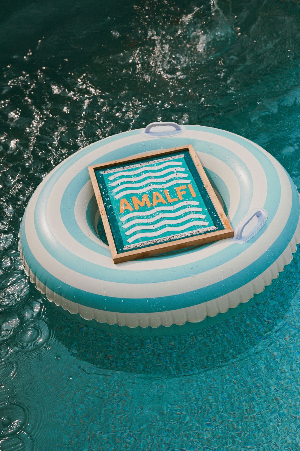 A blue and white striped pool float with an 'Amalfi' sign floats in turquoise swimming pool water.