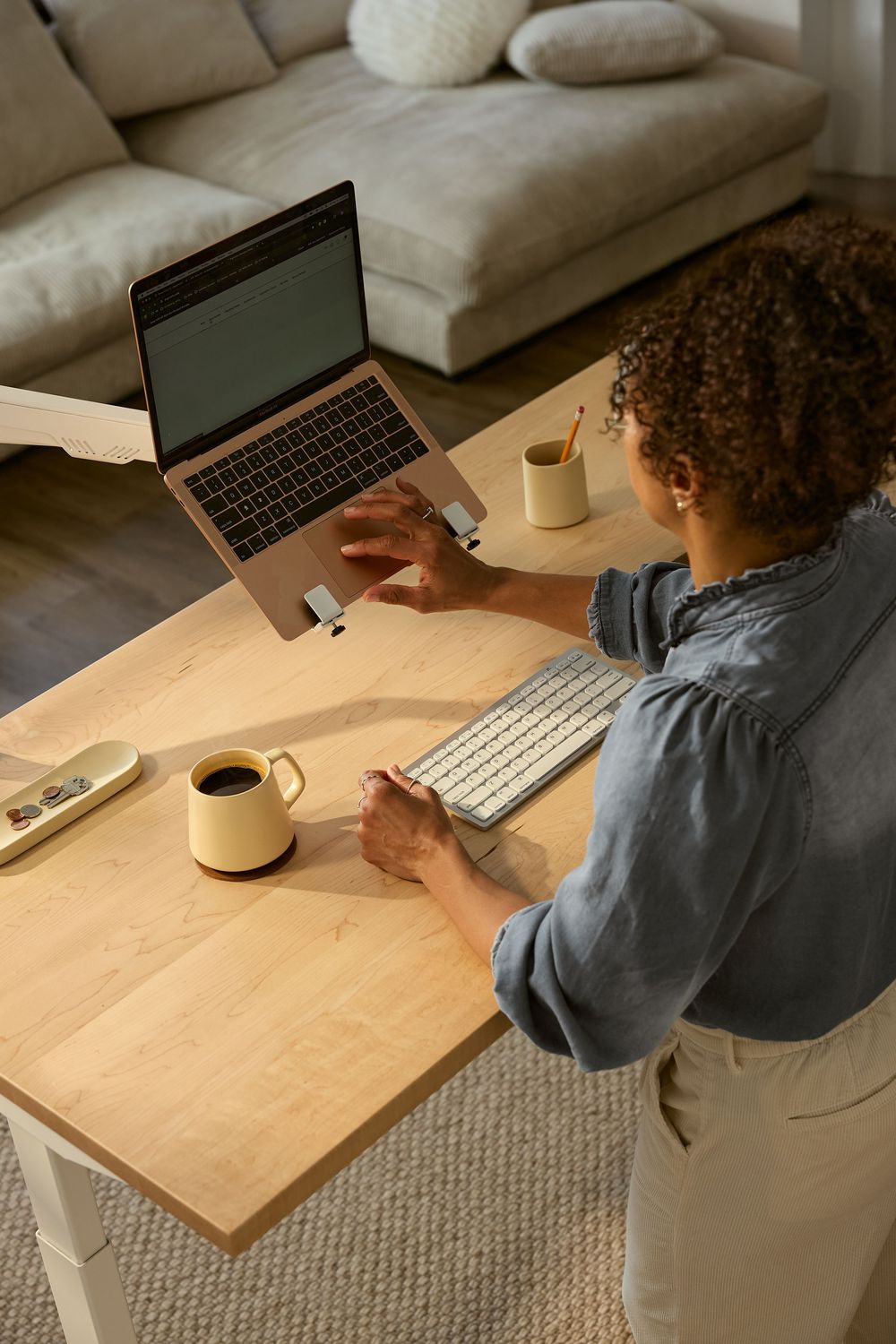 Person working from home at wooden desk with laptop, keyboard and coffee cups in cozy living room setting.