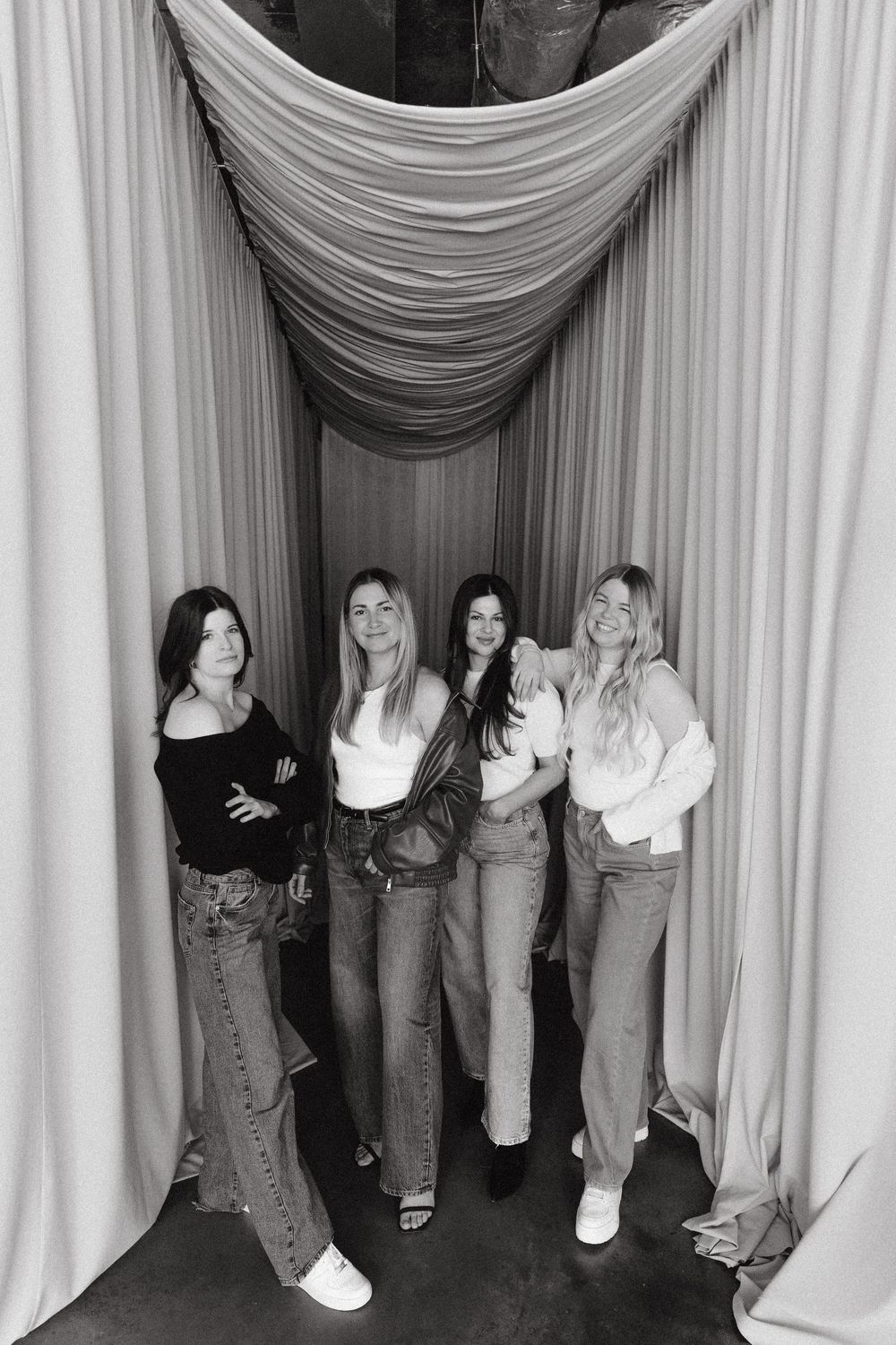 Black and white photograph of four women posing together against white curtains in a studio setting.
