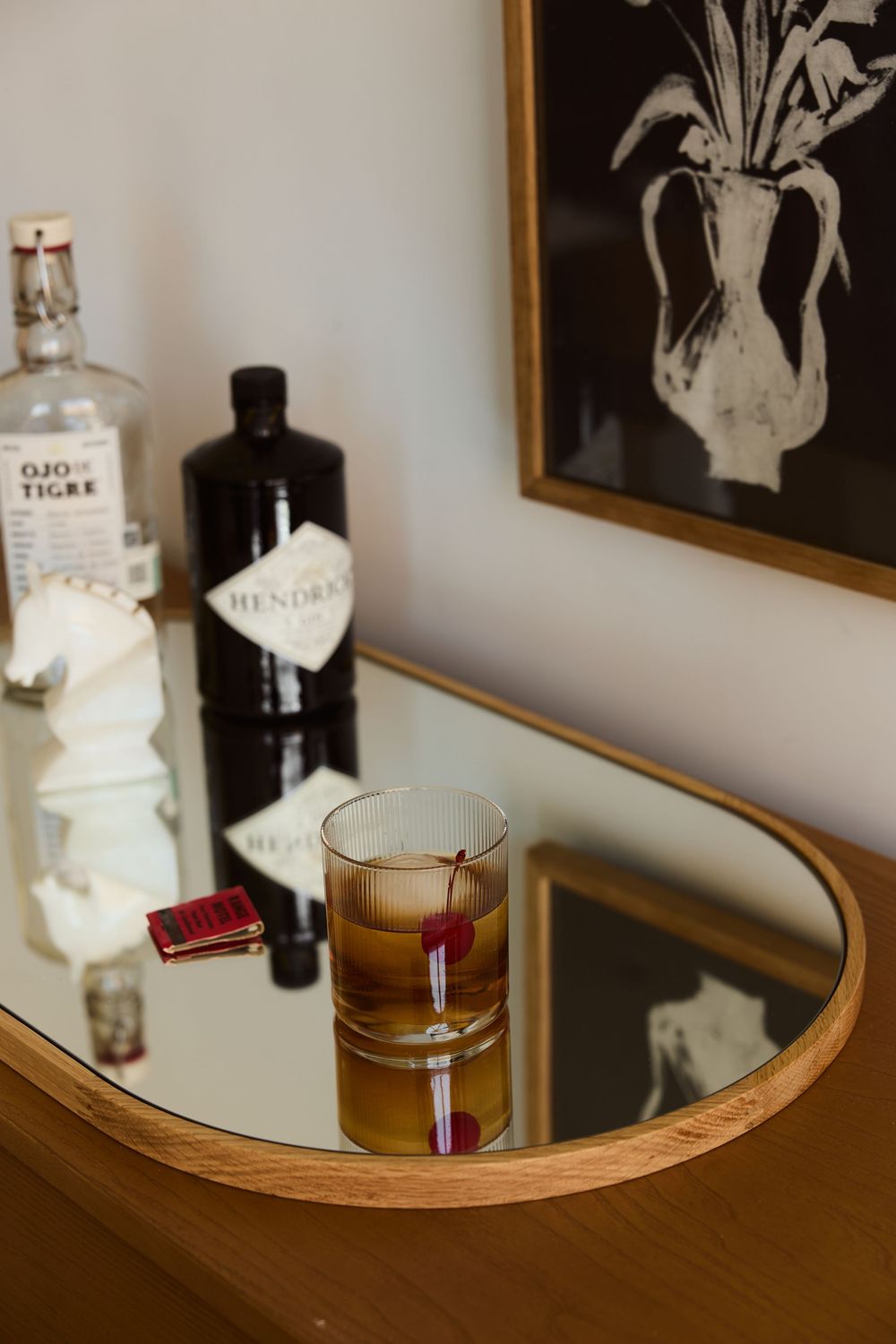A stylish home bar setup with spirits bottles and a craft cocktail on a mirrored surface beneath framed wall art.