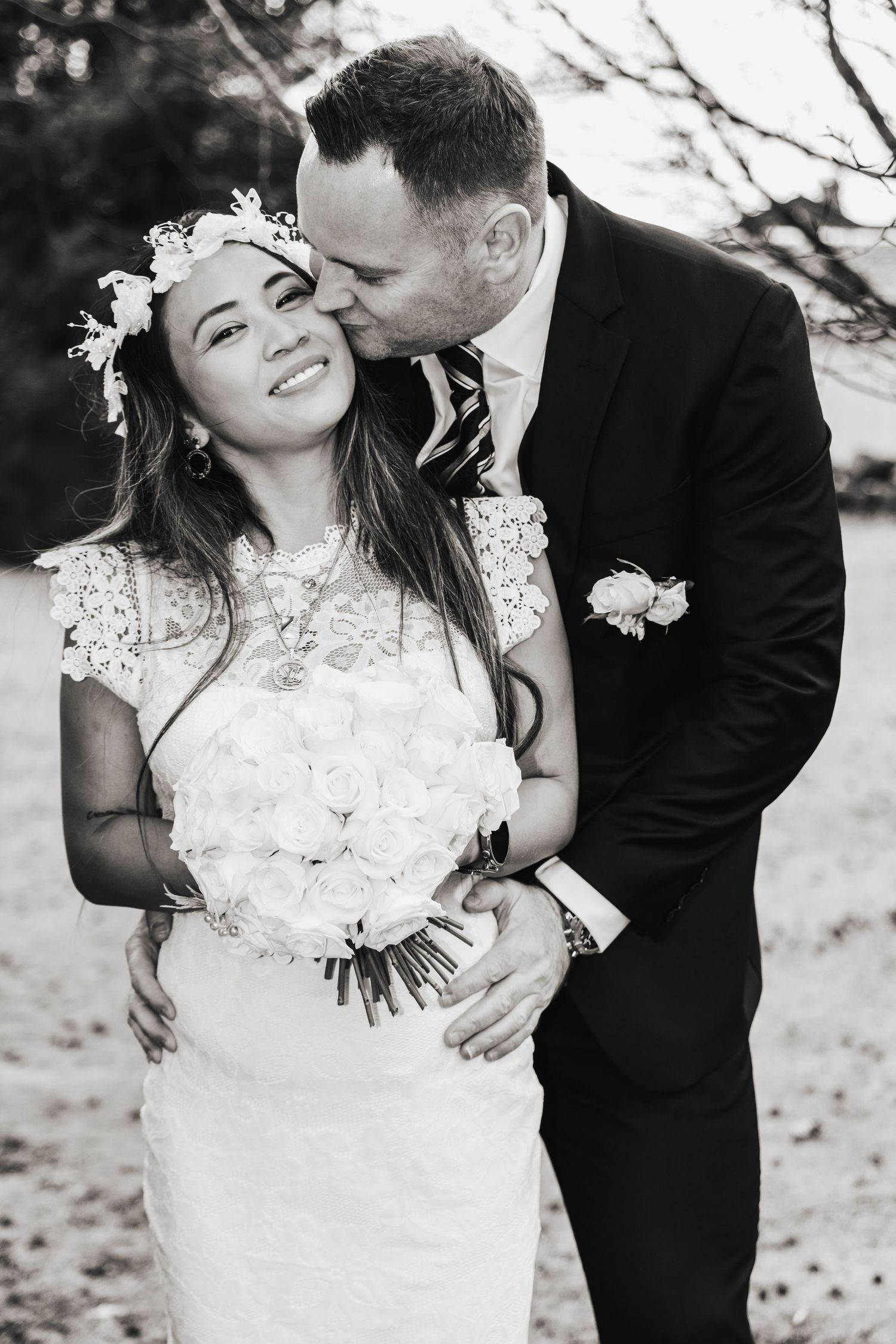 Black and white photo of groom kissing bride wearing floral crown and holding white roses.
