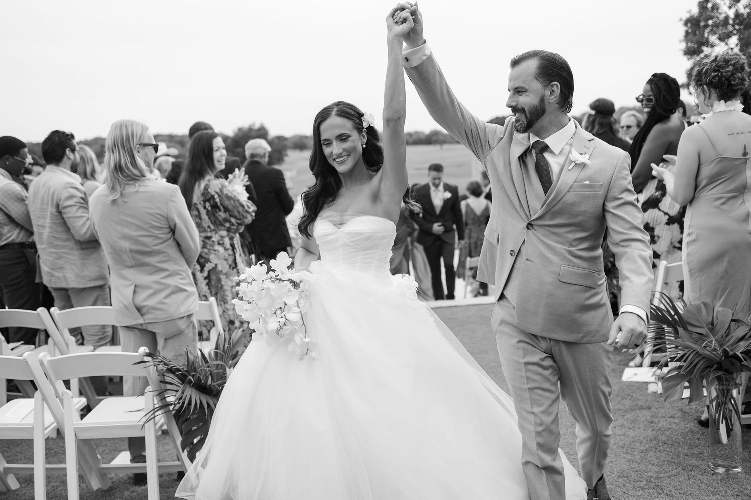 A black and white photo captures a couple dancing joyfully on a hilltop overlooking a lake.