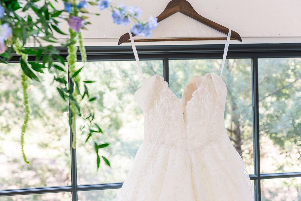 White wedding dress with lace details hangs by window with delicate blue flowers and greenery.