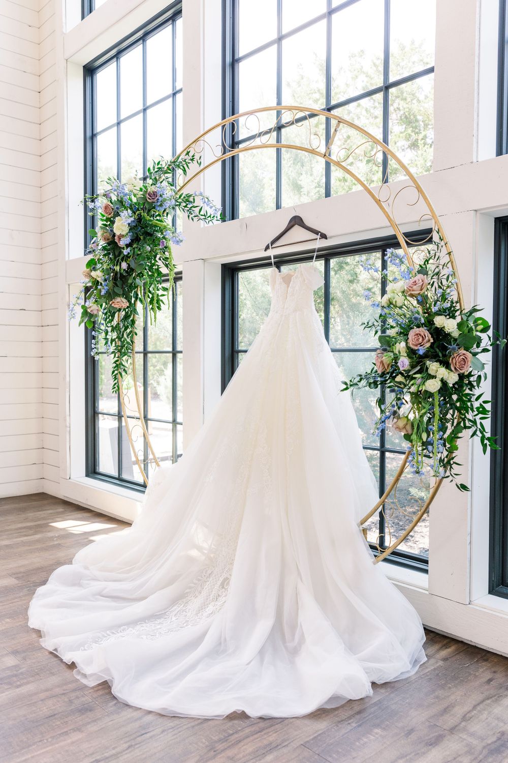 Elegant white wedding gown hanging from a gold arch adorned with pink roses and greenery by sunlit windows.