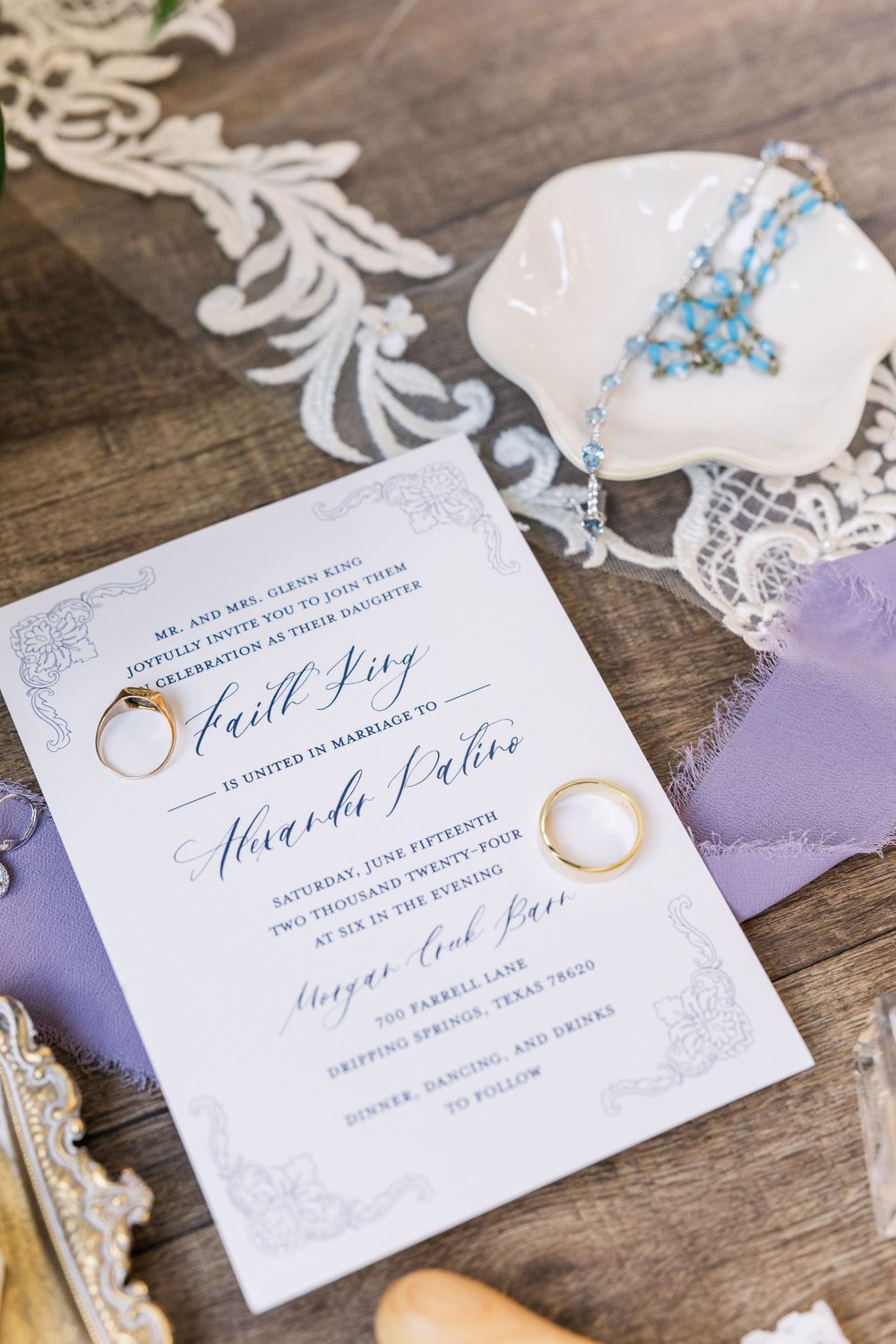 Elegant wedding invitation with gold rings and purple accents displayed on wooden surface with lace details.