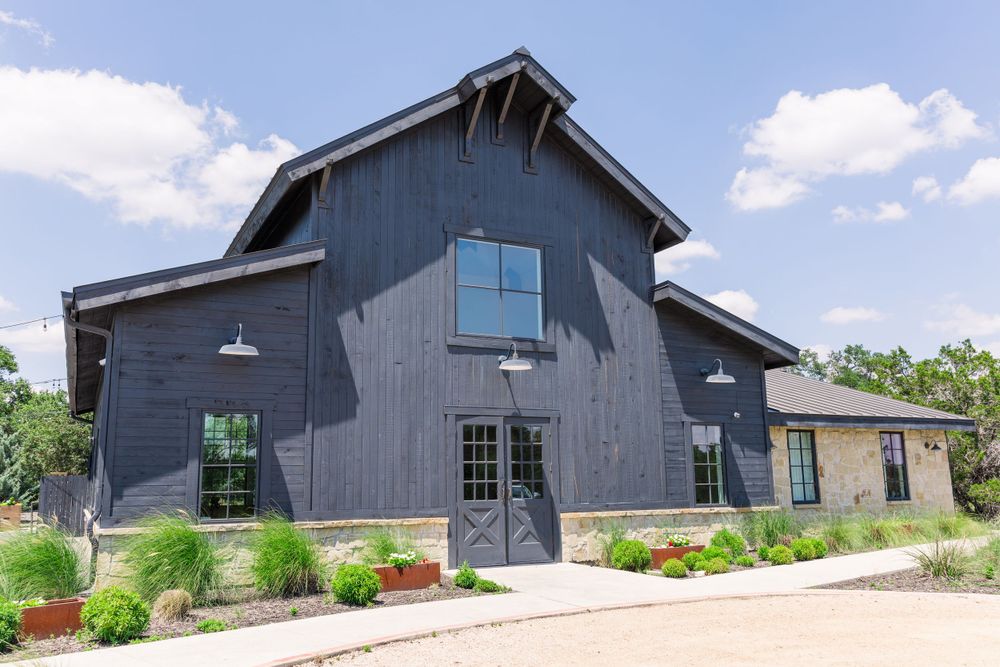 Modern dark gray barn-style event venue with black trim and windows features landscaped garden beds out front.