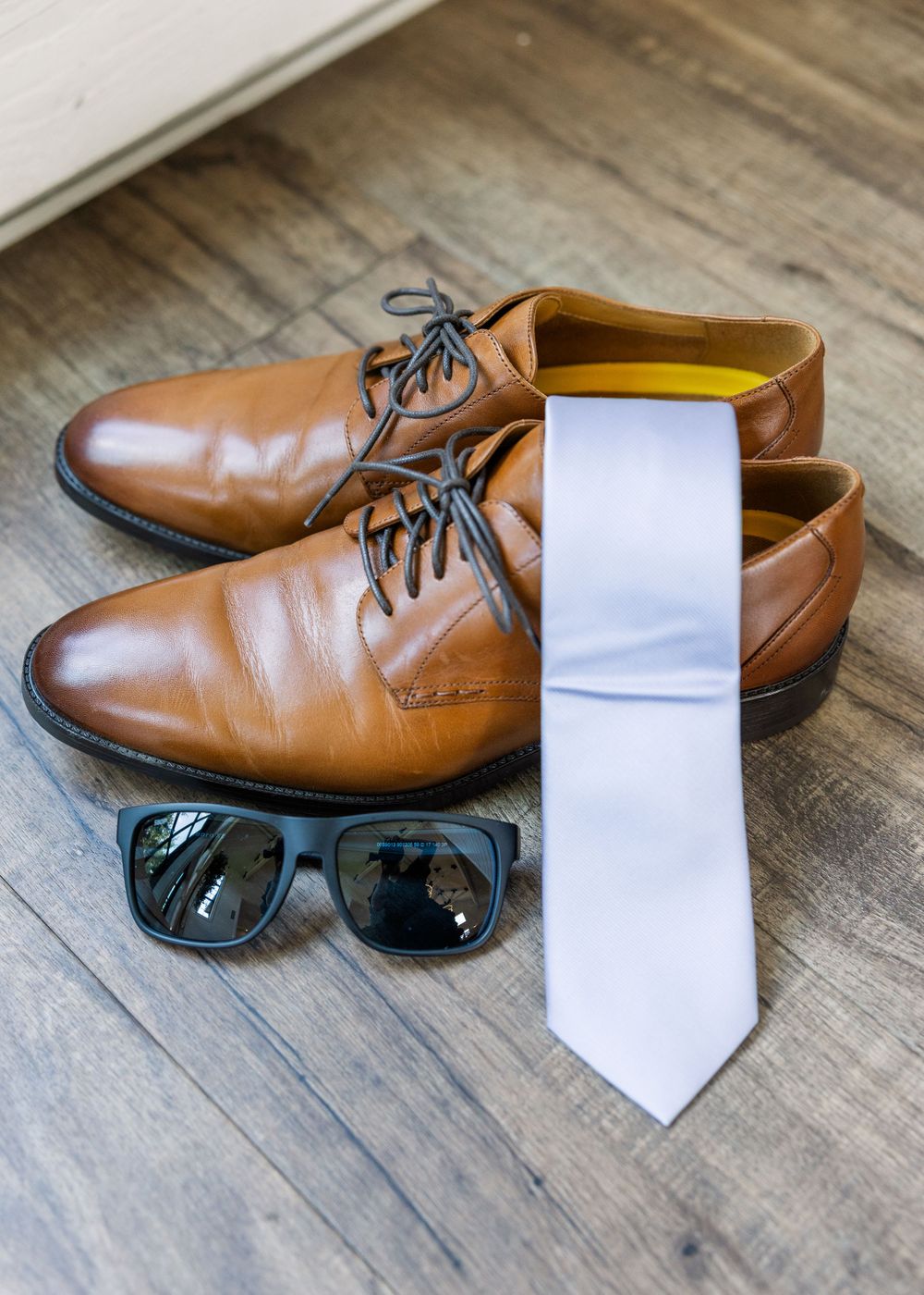 Brown leather dress shoes arranged with a white tie and black sunglasses on a wooden floor.