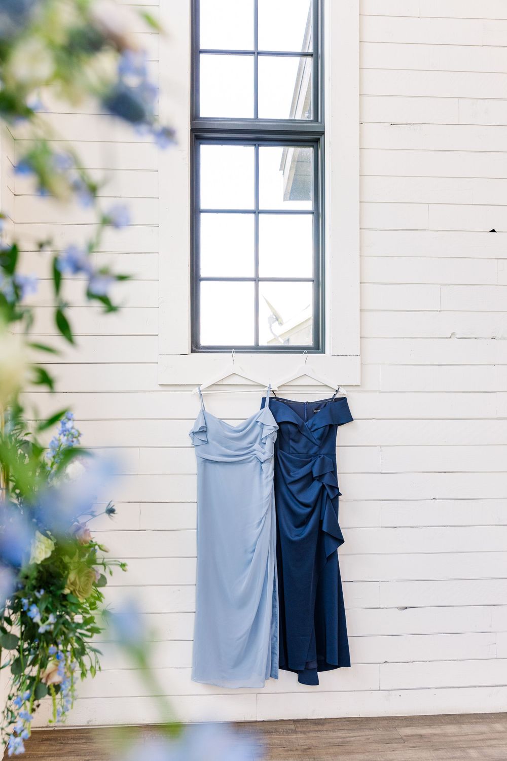 Two bridesmaid dresses in light blue and navy hanging on a white wall near a window with greenery in foreground.