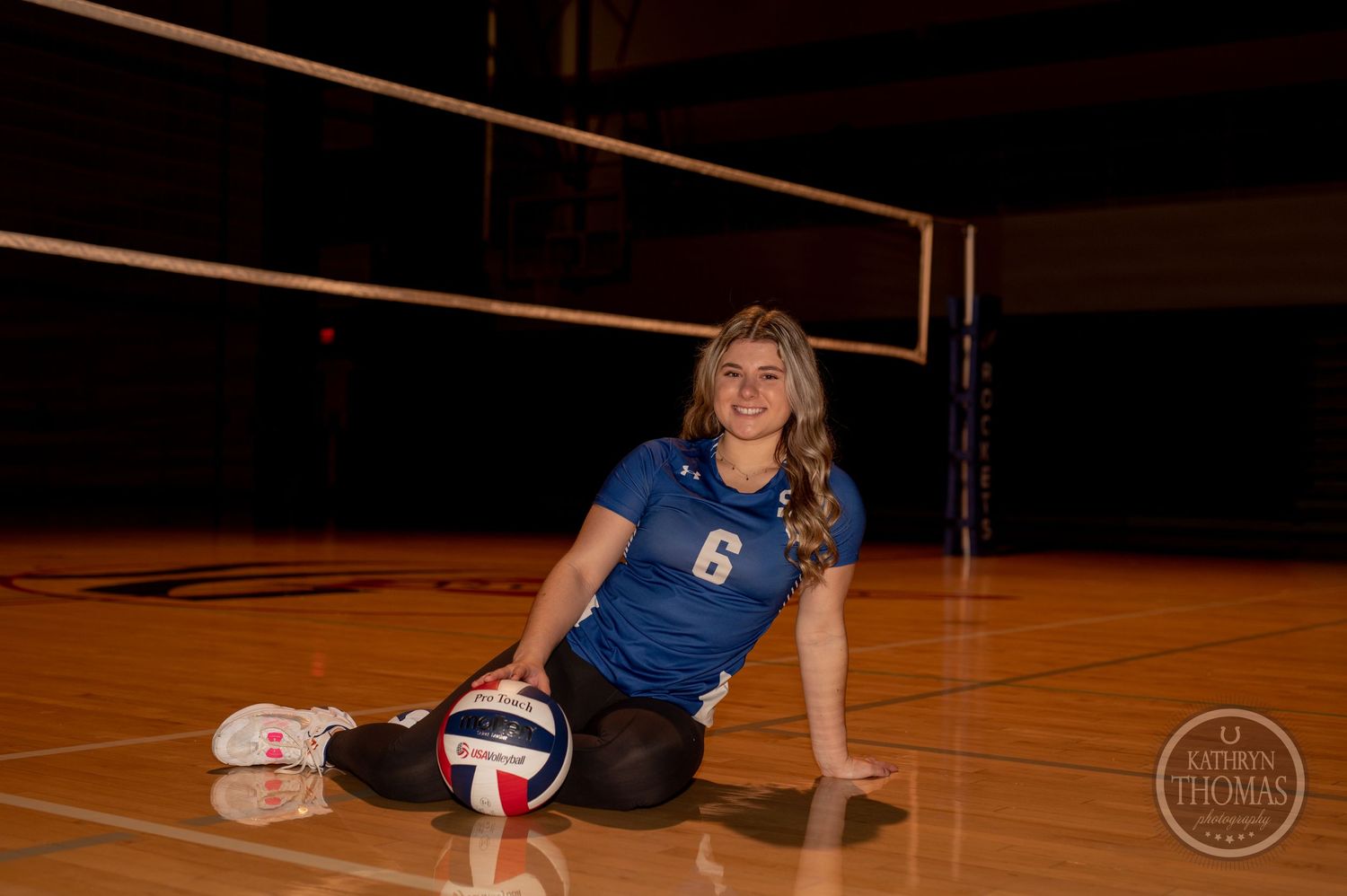 Indoor volleyball player in blue jersey number 6 sitting on court floor with volleyball.
