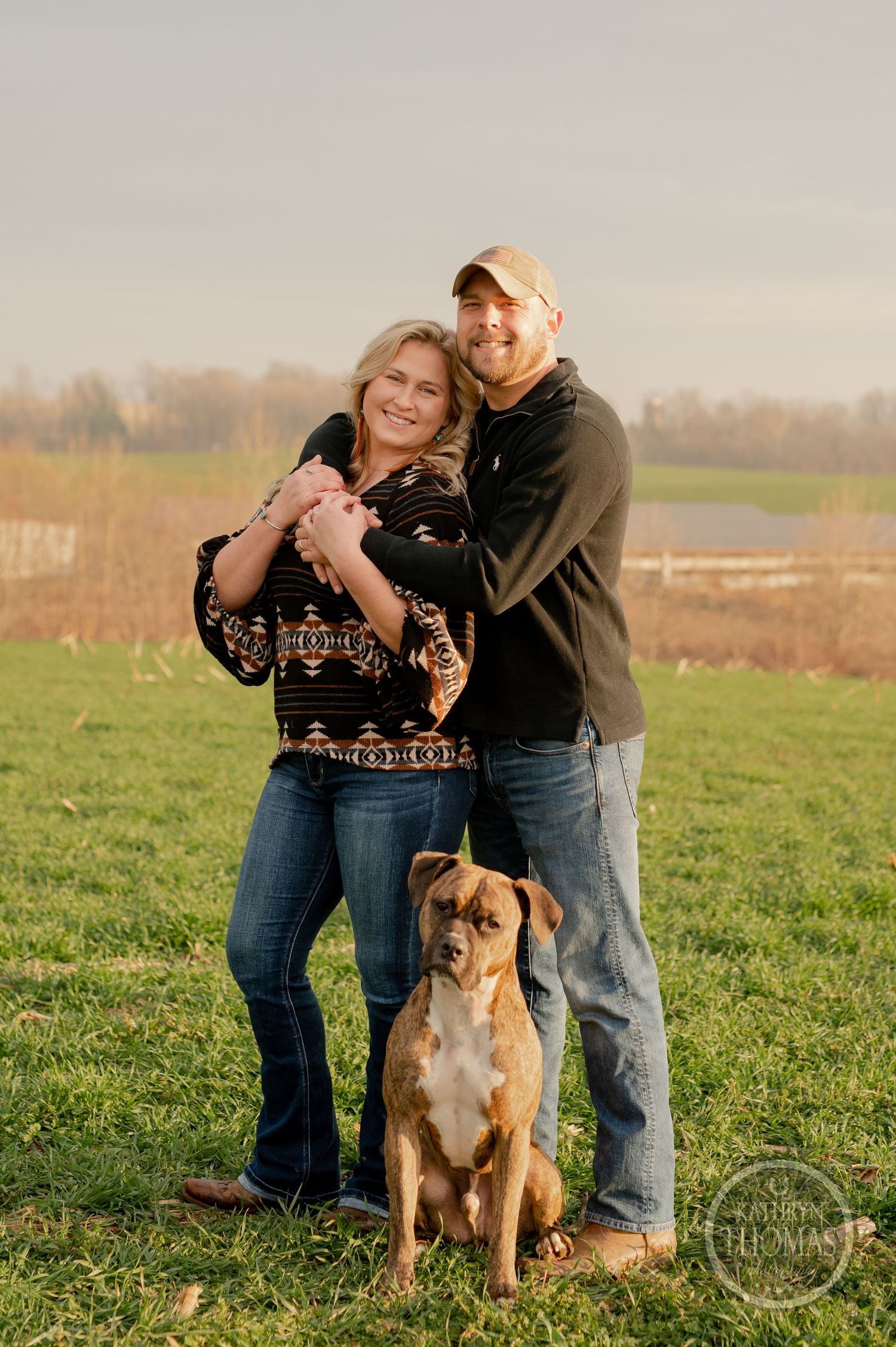 A couple stands in a grassy field with their brown and white pit bull dog at sunset.