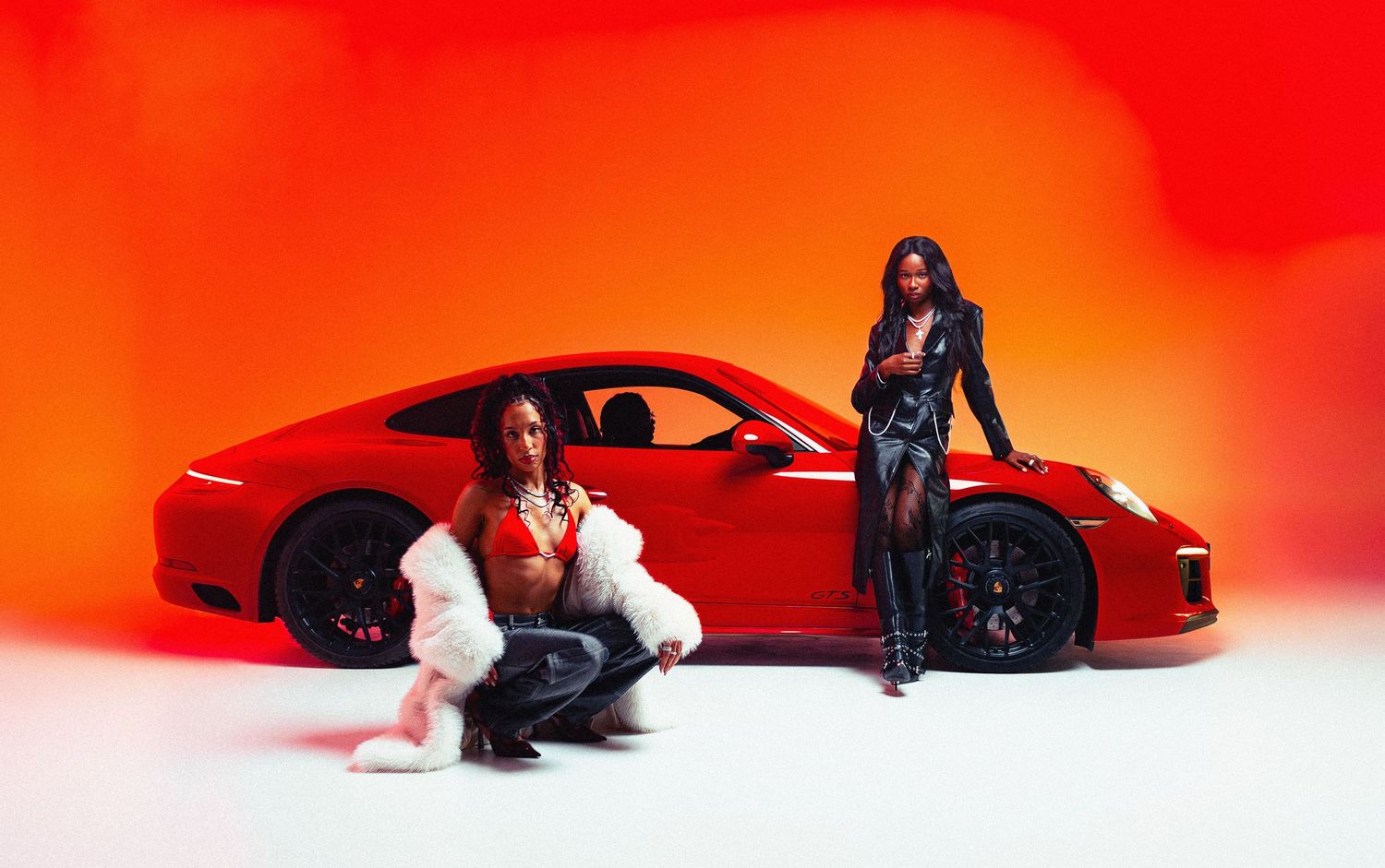 Fashion automotive photo of two models posing with a red Porsche sports car against a vivid orange background