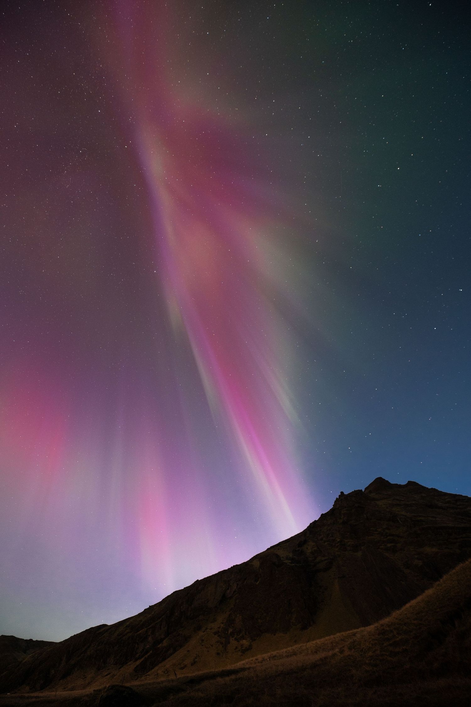 Pink and purple aurora borealis illuminates the night sky over a mountainous silhouette.