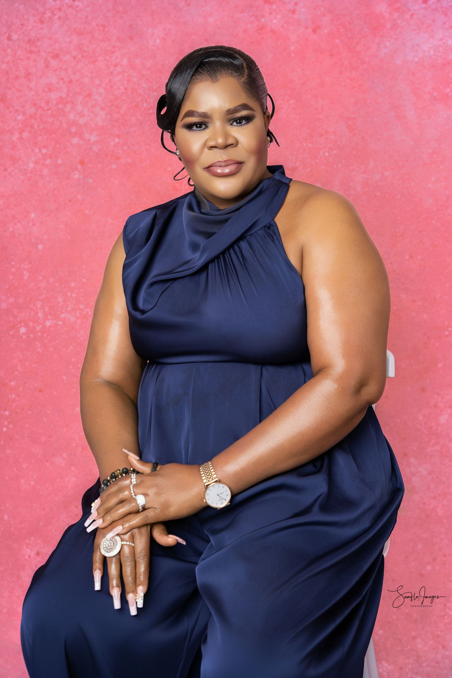 Woman in navy blue satin halter jumpsuit posing confidently against a pink background.