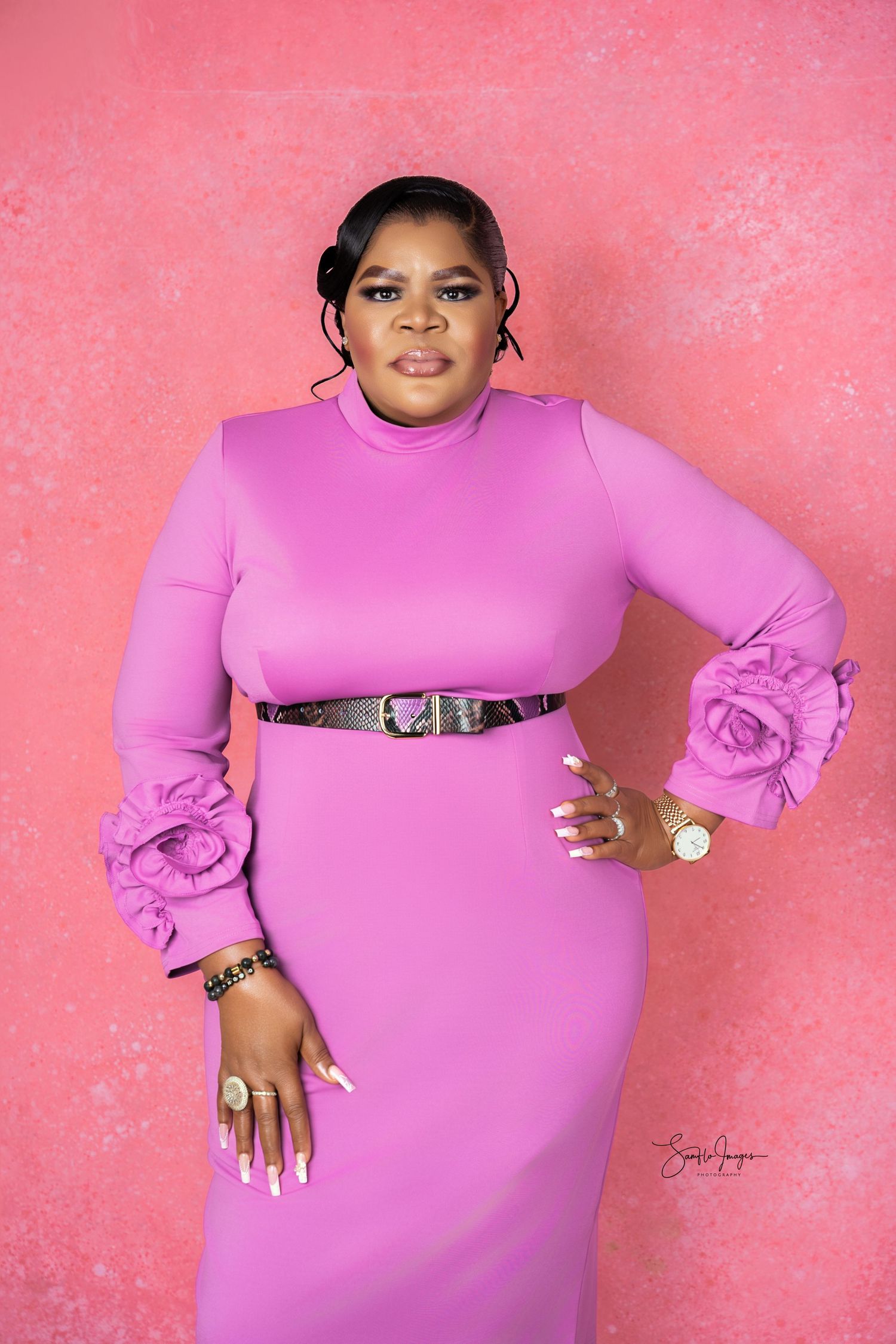Woman in pink midi dress with floral cuffs and black belt poses against pink background.