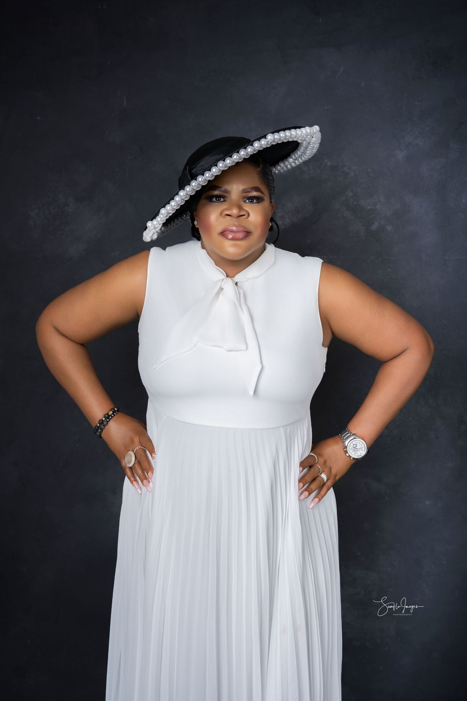 A plus-size woman poses confidently in a white pleated dress and pearl-adorned black hat against a dark studio background.