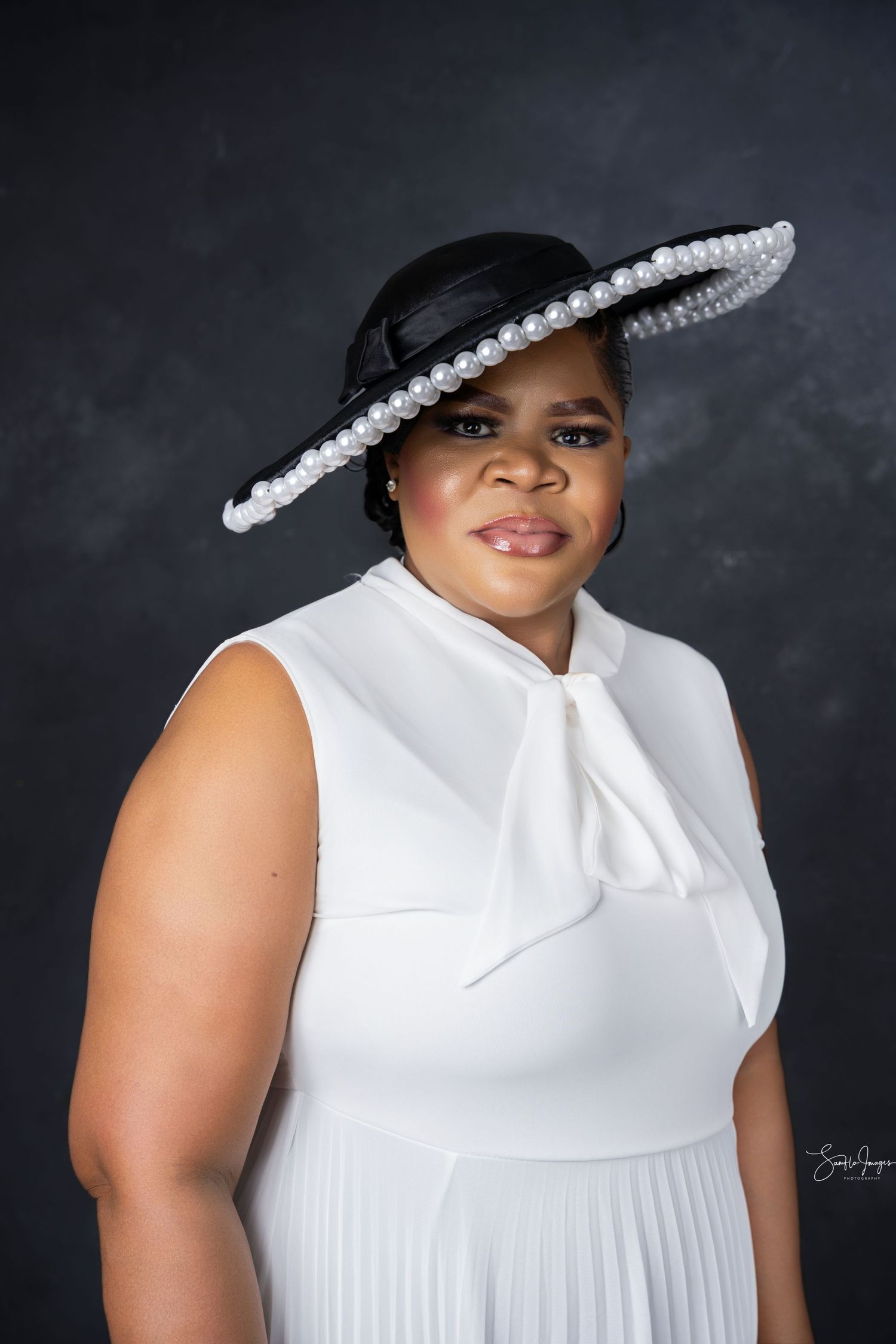 Woman in white sleeveless dress and black wide-brim pearl hat poses for elegant studio portrait.